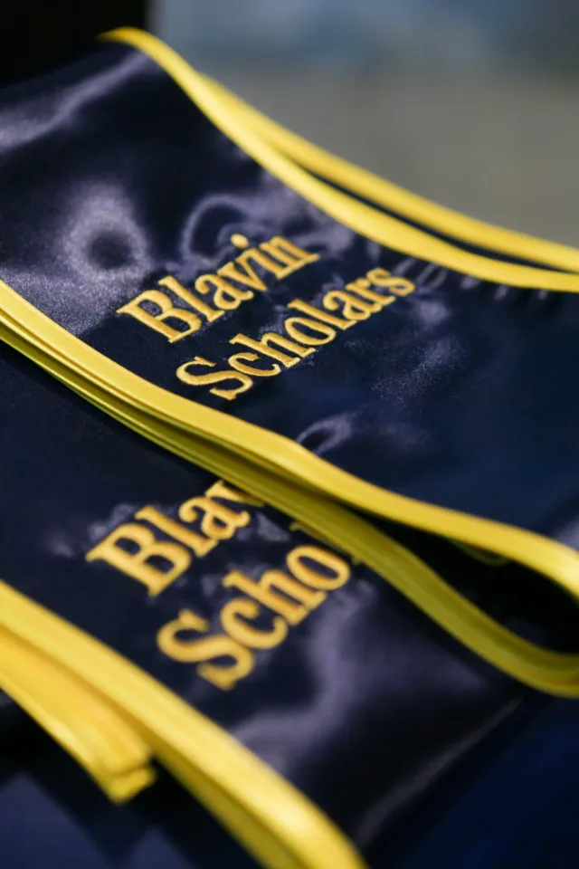 Close-up of folded blue graduation stoles with maize trim, featuring embroidered maize text that reads “Blavin Scholars.”